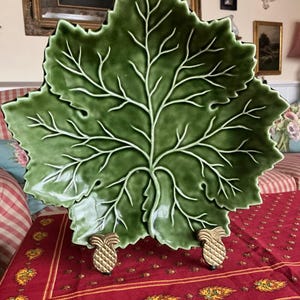 Peut inclure: Un grand plat de service en céramique verte en forme de feuille avec des nervures blanches. Le plat est soutenu par deux pieds dorés en forme d'ananas. Le plat est présenté sur une nappe rouge à motifs floraux.