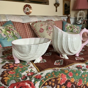 Puede incluir: Un juego de azucarero y jarra de crema de porcelana blanca con detalles rosas y diseño de concha. El juego está sobre un mantel floral.