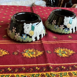May include: Two dark brown ceramic candle holders with a glossy finish. Each piece features a hand-painted white cow with a yellow bell around its neck, surrounded by small green leaves and light blue and pink flowers around the base.