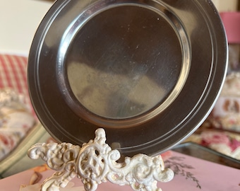 Puede incluir: Un plato de metal plateado con un soporte de metal blanco decorativo y ornamentado. El plato es redondo y tiene un borde elevado. El soporte tiene un diseño de rollo.