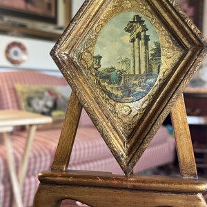 May include: A small, gold-framed picture of an ancient Roman ruin sits on a wooden easel. The picture is set against a background of a striped couch and a patterned rug.
