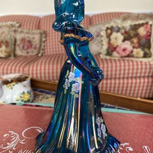 May include: A blue iridescent glass figurine of a woman in a long dress with white flowers painted on it. The dress has gold trim and the figurine is standing on a table.