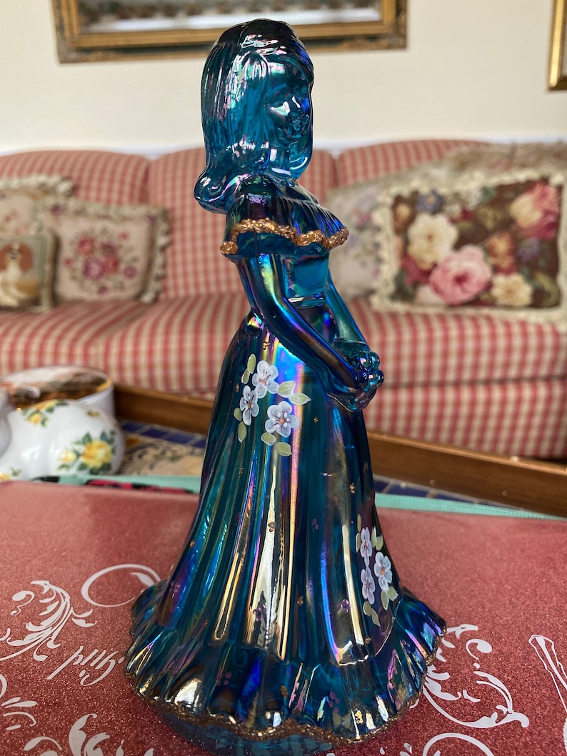 May include: A blue iridescent glass figurine of a woman in a long dress with white flowers painted on it. The dress has gold trim and the figurine is standing on a table.