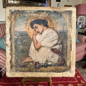 Peut inclure: Une œuvre d'art carrée vintage représentant une figure assise avec un halo doré. La figure porte des vêtements blancs et rouges, avec une expression sereine. L'œuvre d'art a un aspect vieilli et usé, avec une bordure claire et un support rouge et or.