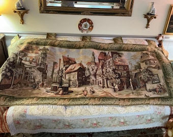Könnte beinhalten: Eine Tapisserie, die einen europäischen Marktplatz mit Gebäuden, einem Brunnen und Bäumen darstellt. Die Tapisserie hat einen grünen und braunen Rand.