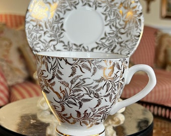 May include: A white teacup and saucer with a gold floral pattern. The cup has a handle and the saucer has a scalloped edge.