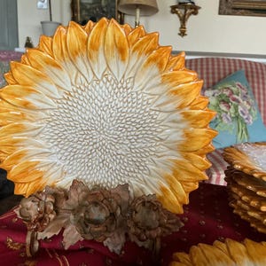May include: A large, decorative ceramic plate in the shape of a sunflower, with a white center and yellow and orange petals. The plate is supported by a brown metal stand with floral accents. Additional plates are stacked in the background.