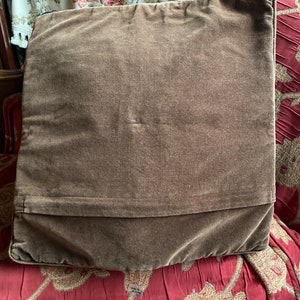 Puede incluir: Una almohada de terciopelo marr&oacute;n con un dise&ntilde;o liso. La almohada est&aacute; sentada sobre una tela de estampado floral rojo y dorado.