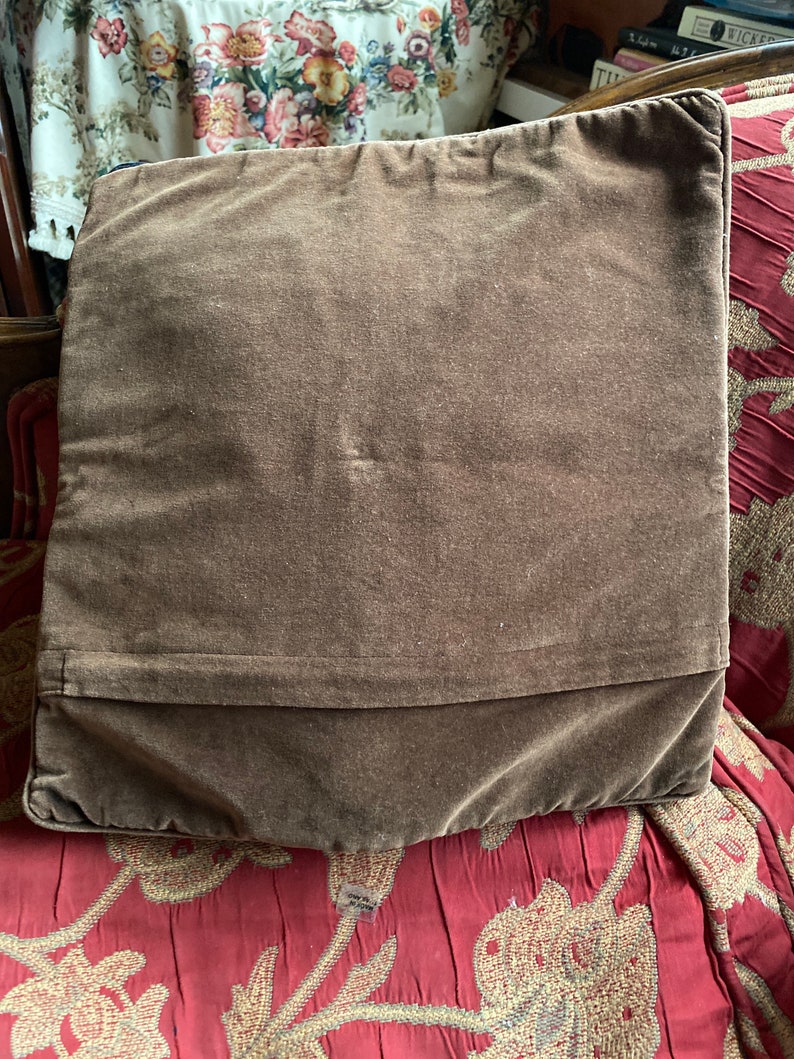 Puede incluir: Una almohada de terciopelo marr&oacute;n con un dise&ntilde;o liso. La almohada est&aacute; sentada sobre una tela de estampado floral rojo y dorado.