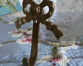 Puede incluir: Un gancho de metal dorado con un diseño de lazo decorativo. El gancho está sobre una tela de estampado azul y blanco.