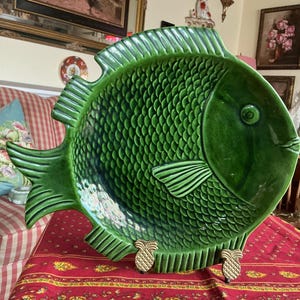 Puede incluir: Una gran fuente de cerámica verde con forma de pez con diseños detallados de escamas y aletas. La fuente está sostenida por dos soportes decorativos dorados con forma de piña. La fuente con forma de pez es una pieza decorativa.