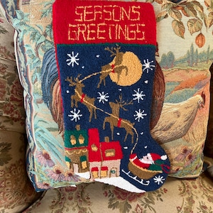 Puede incluir: Una media de Navidad roja y azul con las palabras "Seasons Greetings" bordadas en la parte delantera. La media presenta una escena de renos volando por el cielo nocturno con una luna y estrellas. Hay un pequeño pueblo en la parte inferior de la media y Santa Claus en un trineo.