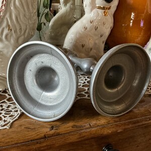 May include: Two antique silver candle holders with a round base and a central stem. The candle holders are made of metal and have a worn, vintage appearance. The background includes decorative porcelain figurines.