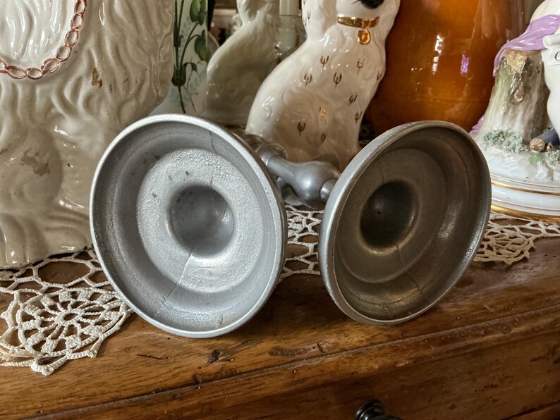 May include: Two antique silver candle holders with a round base and a central stem. The candle holders are made of metal and have a worn, vintage appearance. The background includes decorative porcelain figurines.
