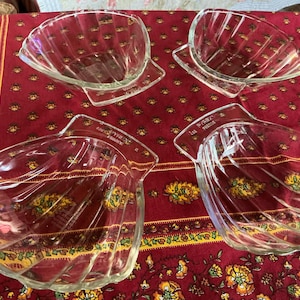 Op de afbeelding: Vier heldere glazen serveerschalen met een schelpontwerp. Elke kom heeft een uitlopend, geribbeld interieur en een rechthoekige basis. De kommen zijn gerangschikt op een rood patroon tafelkleed.