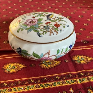 May include: A white ceramic trinket box with a lid, featuring a floral and bird design in pink, blue, green, and orange. The box has a gold rim and sits on a red patterned tablecloth.