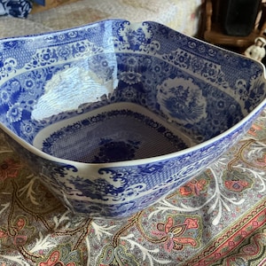 May include: A large, square ceramic bowl featuring a classic blue and white transferware pattern. The design includes intricate floral motifs, geometric borders, and detailed landscape scenes with pagodas, all rendered in a deep cobalt blue.