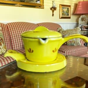 Puede incluir: Una tetera de cerámica amarilla con tapa y asa, sentada sobre un platillo amarillo a juego. La tetera tiene un diseño floral en el lateral.