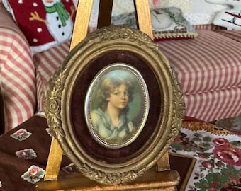 Puede incluir: Un retrato ovalado en oro de una joven con cabello castaño claro y un vestido azul. El retrato está enmarcado en oro y colocado sobre un fondo de terciopelo burdeos. Se muestra en un caballete de madera.