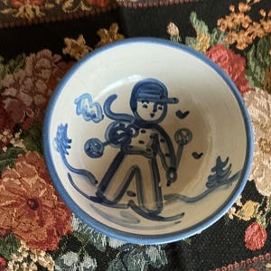 May include: A small, round ceramic bowl with a white base and a blue rim. The interior features a hand-painted blue illustration of a person skiing, wearing a hat and holding ski poles, surrounded by simple blue pine tree and heart motifs.