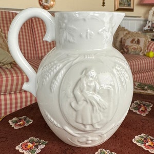May include: White ceramic pitcher with a raised design of a woman holding a sheaf of wheat. The pitcher has a wide, round body and a handle that curves up and over the top.