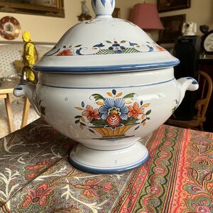 Może przedstawiać: Biała ceramiczna waza z pasującą pokrywką, ozdobiona ręcznie malowanymi motywami kwiatowymi w kolorach niebieskim, pomarańczowym i zielonym. Misa posiada dwa boczne uchwyty, podstawę oraz szpiczasty uchwyt pokrywki, wykończone niebieskimi liniami.