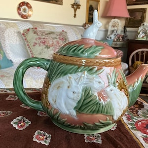 Può includere: Una teiera in ceramica con una glassa rosa e verde, con un design di conigli bianchi e viti con foglie. La teiera ha un coperchio con una statuetta di coniglio bianco in cima.