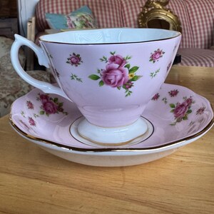 May include: A pink teacup and saucer set with a floral rose design. The teacup has a white handle and a gold trim. The saucer also has a gold trim. The set is on a wooden table.