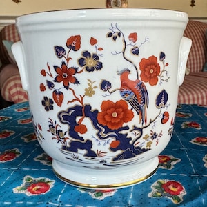 May include: White ceramic planter with a blue, gold, and red floral design featuring a bird. The planter has two handles on the sides.
