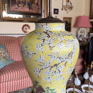 Peut inclure: Une base de lampe en porcelaine jaune avec un motif floral de fleurs blanches et de feuilles vertes. La base de la lampe est sur un piédestal en bois noir avec un motif sculpté. La lampe a deux luminaires en laiton avec des cordons blancs.