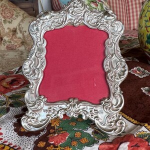 May include: A silver picture frame with ornate details and a red velvet backing. The frame is sitting on a floral patterned fabric.
