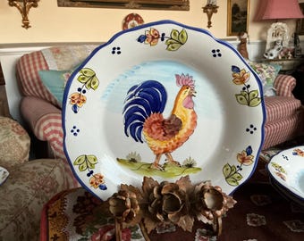 May include: A white ceramic plate with a blue rim features a hand-painted design of a rooster with a blue tail and red and yellow feathers. The plate has a blue and white floral pattern around the edge.