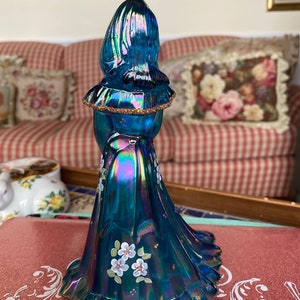 May include: A blue iridescent glass figurine of a woman wearing a long dress with white flowers painted on it. The dress has a ruffled hem and the woman's hair is pulled back.