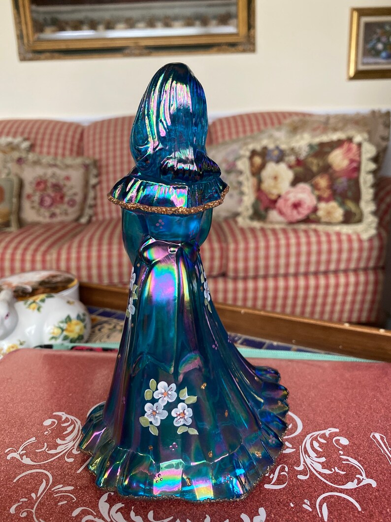 May include: A blue iridescent glass figurine of a woman wearing a long dress with white flowers painted on it. The dress has a ruffled hem and the woman's hair is pulled back.