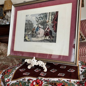 May include: A framed antique print of a scene with people in a garden setting. The print is in black and white and has a gold frame. The frame is on a stand with a floral pattern.