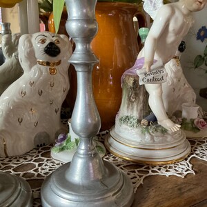 May include: A silver-colored candlestick with a wide base and a slender, ornate stem. The candlestick is made of metal and has a matte finish. The background includes decorative figurines and a vase with flowers.