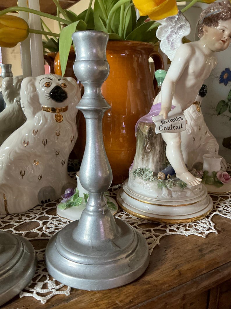 May include: A silver-colored candlestick with a wide base and a slender, ornate stem. The candlestick is made of metal and has a matte finish. The background includes decorative figurines and a vase with flowers.