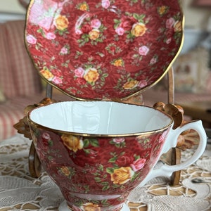 Puede incluir: Un juego de taza y platillo de té vintage. El juego presenta un fondo rojo oscuro con un patrón floral de rosas amarillas y rosas. La taza tiene un interior blanco y un borde dorado. El platillo también es rojo con el mismo diseño floral y ribete dorado.