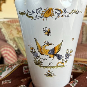May include: White ceramic vase with a scalloped rim and a floral design featuring a yellow bird with blue accents. The vase is decorated with green, yellow, and blue flowers.