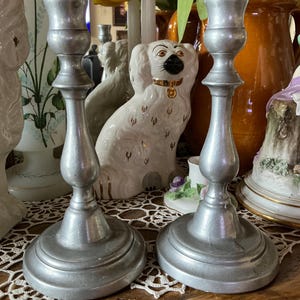 May include: Two silver-colored metal candlesticks with a classic design. The candlesticks have a round base, a slender stem, and a cup-shaped top to hold candles. The background includes decorative items like a ceramic dog figurine and vases.