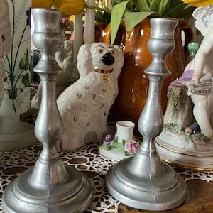 May include: Two silver-colored metal candlesticks with a classic design. The candlesticks have a round base, a central stem, and a top for holding candles. The background includes decorative items like a ceramic dog figurine and a vase of flowers.
