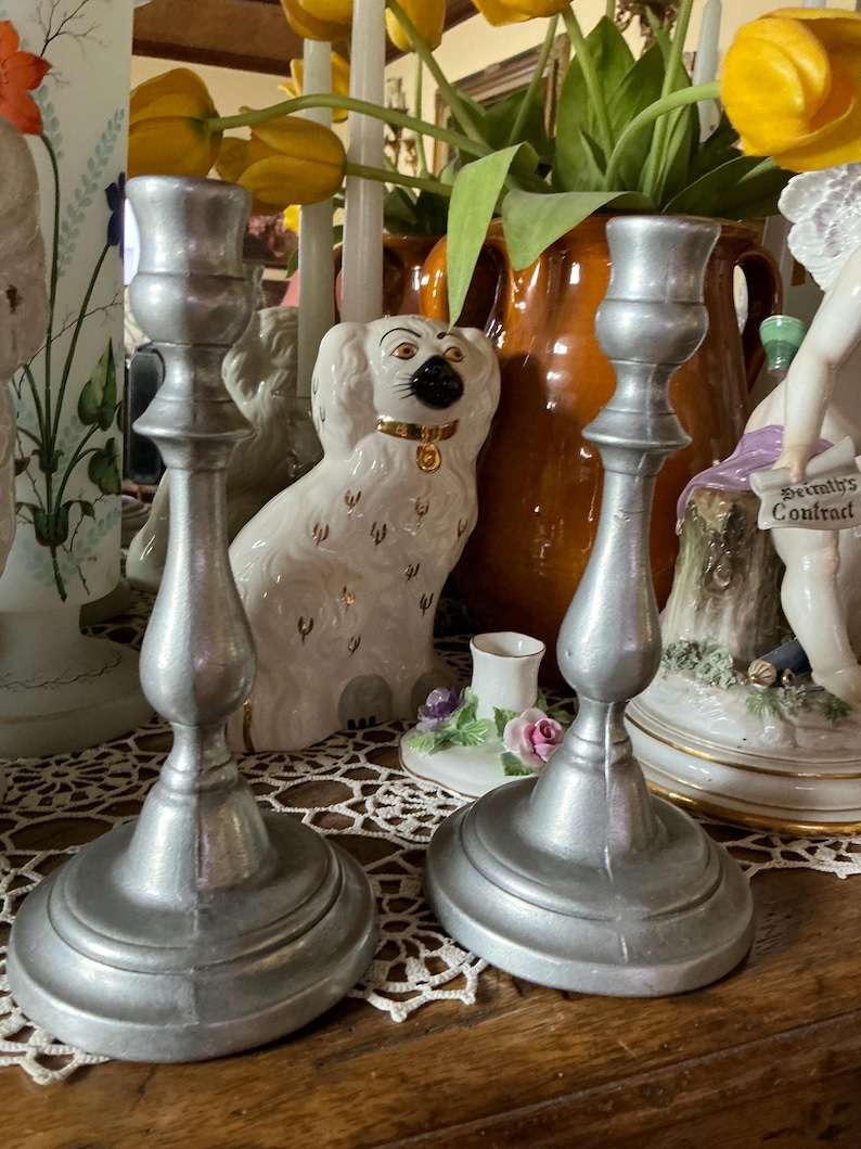 May include: Two silver-colored metal candlesticks with a classic design. The candlesticks have a round base, a central stem, and a top for holding candles. The background includes decorative items like a ceramic dog figurine and a vase of flowers.