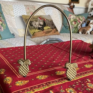 May include: A gold-colored metal plate holder with pineapple-shaped accents. The arched handle is supported by two pineapple designs. The plate holder is on a red tablecloth with a floral pattern.