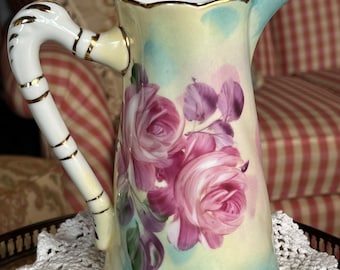 May include: A vintage porcelain pitcher with a white handle and gold trim. The pitcher features a floral design with pink roses and purple leaves on a light green and blue background.