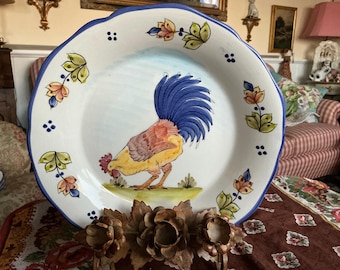 May include: A white ceramic plate with a blue rim and a hand-painted design of a rooster with a blue, red, and yellow tail feathers. The plate is decorated with blue dots and green and yellow floral designs.