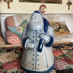 May include: A ceramic Santa Claus figurine featuring a light grey coat with dark blue trim, a matching blue hat, and a blue sack slung over one shoulder. The figure has a smooth, glossy finish with painted blue buttons and vertical line details.