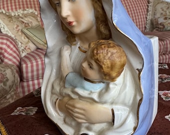 Op de afbeelding: Een wit porseleinen beeldje van de Maagd Maria die een baby Jezus vasthoudt. Het beeldje is geschilderd met blauwe en gouden accenten en heeft een halo met blauwe sterren.