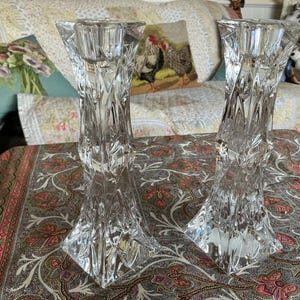 May include: A pair of clear, faceted crystal candle holders featuring a geometric, multi-sided design that tapers towards the centre. Each holder has a circular top opening for a candle and a wide, stable base with intricate, reflective glass cuts.