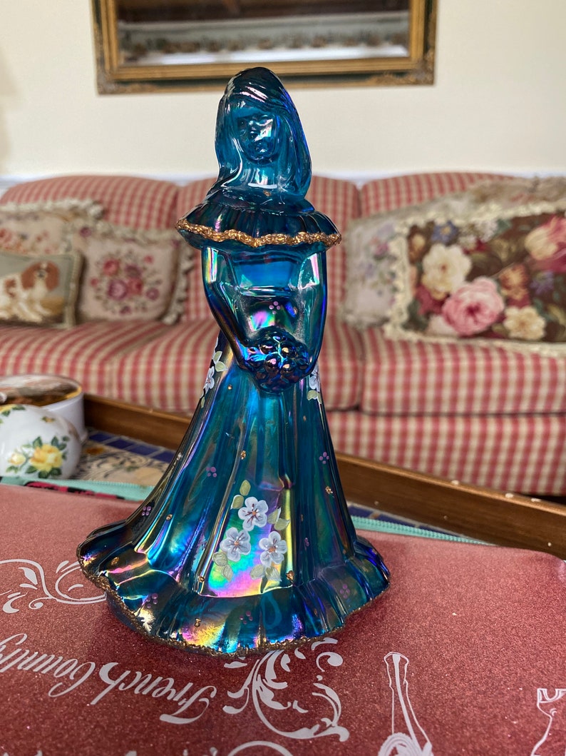 May include: A blue glass figurine of a woman in a long dress with white flowers. The figurine has a gold trim and iridescent finish.