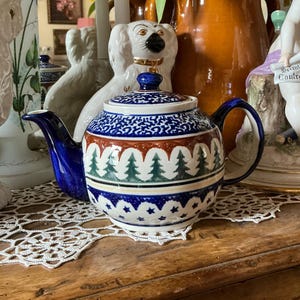 Op de afbeelding: Een keramische theepot met een blauw deksel en handvat, met een decoratief patroon van bomen, sterren en geometrische vormen in blauw, rood en groen. De theepot staat op een houten oppervlak.